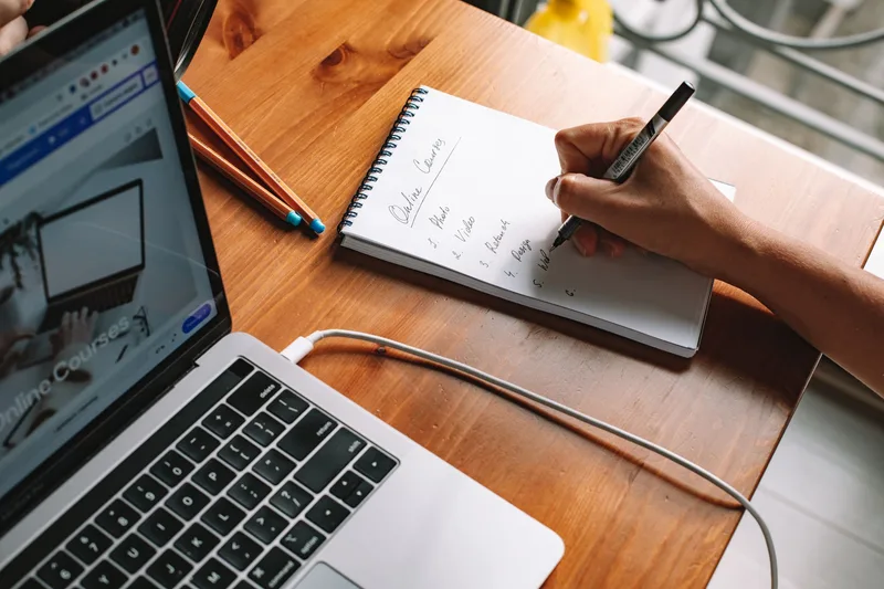 Person writing notes with a laptop open, researching course platform options